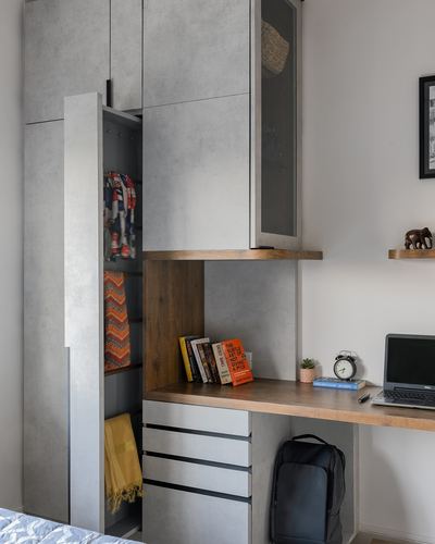 Grey wardrobe with open door beside a wood-topped study desk with laptop and books
