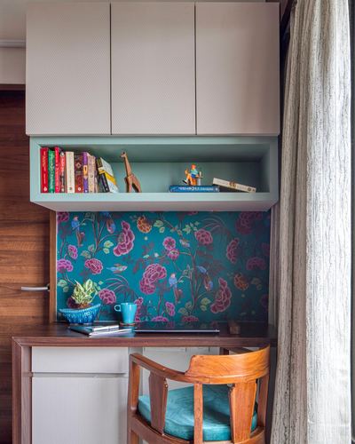 Study nook with floral teal wallpaper, wooden desk, chair, and open bookshelf with books and figurines