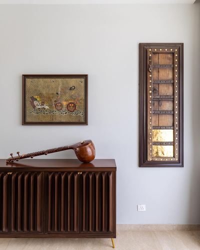 Wall with framed folk painting and antique door-panel mirror above a fluted wood sideboard holding a sarod