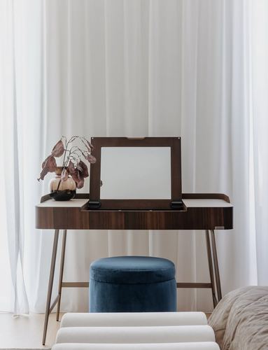 Wood dressing table with flip-up mirror, vase of dried branches, and blue velvet pouf against sheer curtains