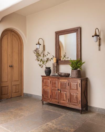Entryway with arched wood door, carved mirror, wooden cabinet, stone floor, brass sconces
