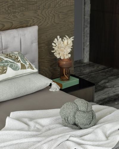 Bed with grey velvet headboard, knit cushions, and dried floral vase against wood-panelled wall