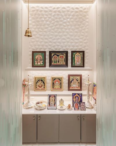 Pooja unit niche with tanjore paintings, brass bell, and silver lamps on a textured wall