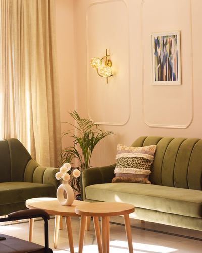 Olive green velvet sofa and armchair in a sunlit blush pink living room with brass sconce and wooden nesting tables