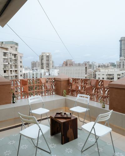 Terrace corner with white folding chairs around wooden stool, terracotta jali railing, city skyline behind