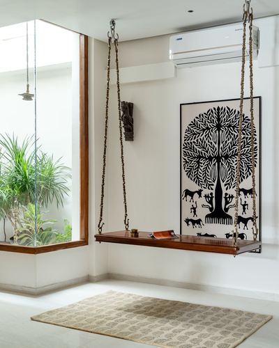 Wooden swing bench hangs by brass chains beside a black and white warli tree painting