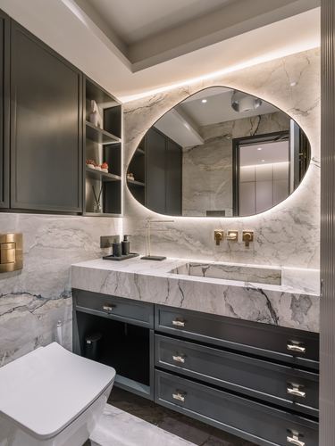Bathroom vanity with marble walls, backlit oval mirror, brass fixtures and dark cabinetry