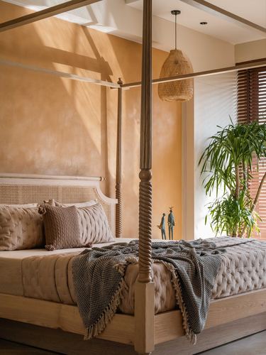 Four-poster wooden bed with cane headboard against a terracotta plaster wall, sunlight streaming in beside a woven pendant light.