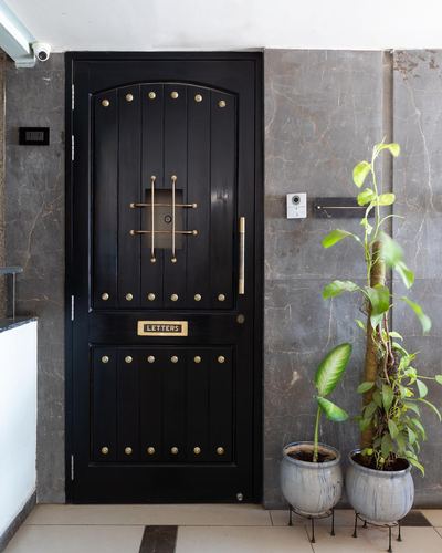 Black studded wooden entry door with brass grille and letterbox, beside stone wall and potted plants