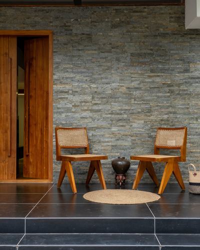 Two teak and cane chairs flank a metal stool on a black stone porch against a textured stone wall