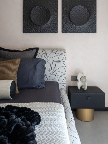 Bedroom corner with black textured wall art, patterned headboard, dark cushions, and a black nightstand on brass base
