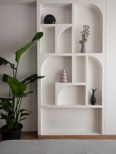 White sculptural shelving unit with arched niches holding ceramic vases beside a large potted plant