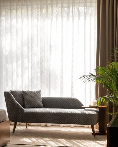Grey patterned chaise lounge in front of sheer curtains with soft daylight and a potted palm