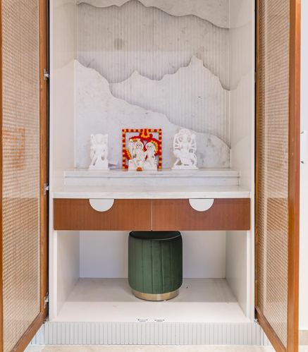 Pooja cabinet with marble cloud-pattern backdrop, white idols, and green velvet stool below, flanked by cane doors