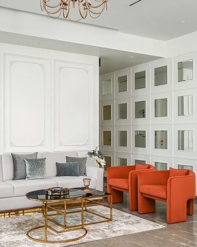 White neo-classical living room with mirrored panel wall, grey sofa and two orange armchairs around a brass coffee table