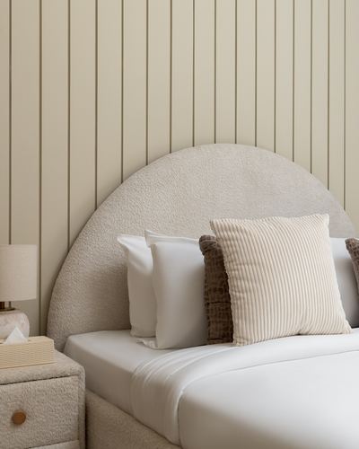 Arched boucle headboard with layered cushions against vertical wood panelling, bedside lamp and nightstand