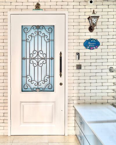 White paneled door with ornamental iron scrollwork grille, set in a brick-pattern tiled wall with a lantern sconce.