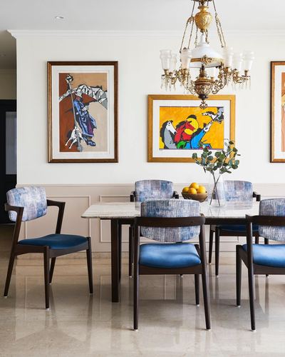 Dining room with blue upholstered chairs, marble table, brass chandelier, and colorful framed art on wall