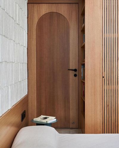 Arched wood door beside textured white wall and fluted wood partition with bookshelf, bed in foreground