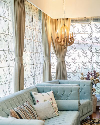 Pale blue tufted sofa beneath a wooden chandelier, framed by leaf-print roman blinds and cream curtains.