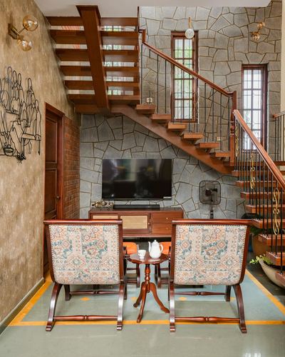 Double-height living room with wooden staircase against stone wall, TV console below, two patterned armchairs and round table in foreground