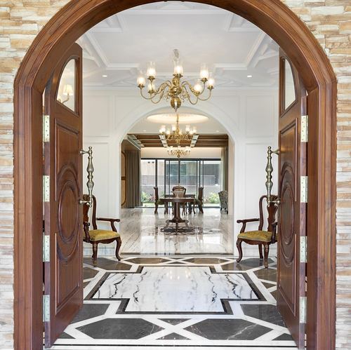 View through open carved wooden arched doors into a marble-floored foyer with chandeliers and a dining area beyond