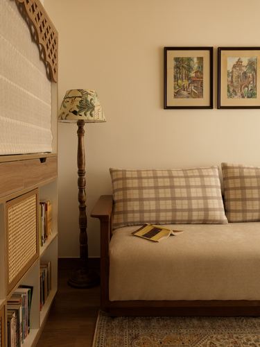 Beige sofa with checkered cushions beside a floor lamp and cane bookshelf under framed art