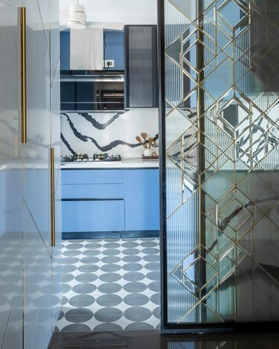 View through brass-framed glass partition into a blue kitchen with patterned tile floor
