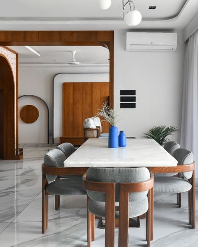 Marble dining table with grey upholstered chairs beneath pendant lights, arched wood portal to living area beyond