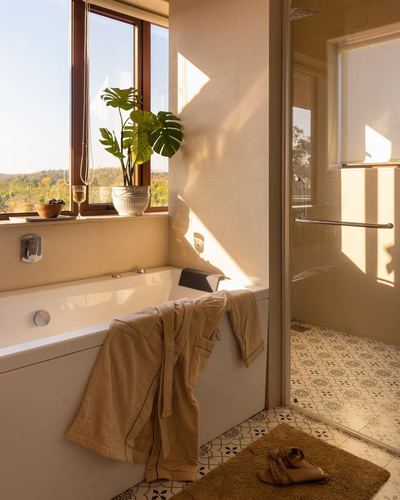 Sunlit bathroom with soaking tub, window view of green hills, and patterned tile shower floor