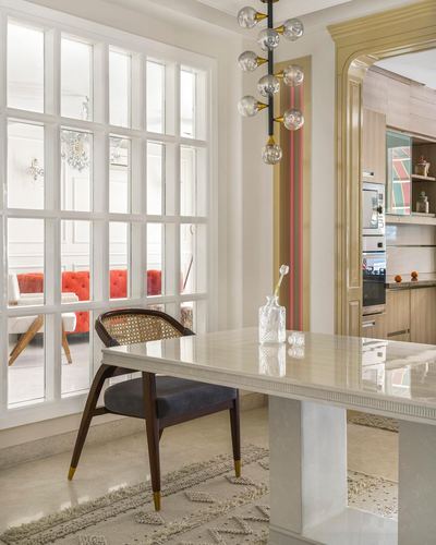 Marble dining table with cane-back chair beside a large grid window, kitchen visible through gold-trimmed archway