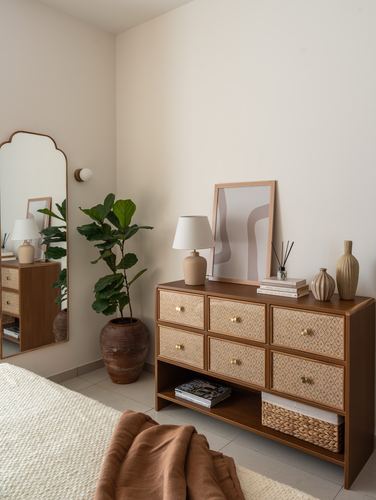 Sunlit bedroom corner with cane-front wood dresser, arched mirror, and fiddle leaf fig plant