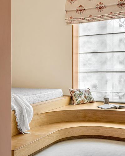 Curved wooden window seat with floral cushion, throw blanket, and sheer curtain light