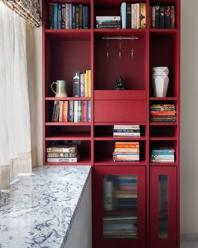 Red built-in bookshelf with books, brass bells, and a small idol beside a marble window seat