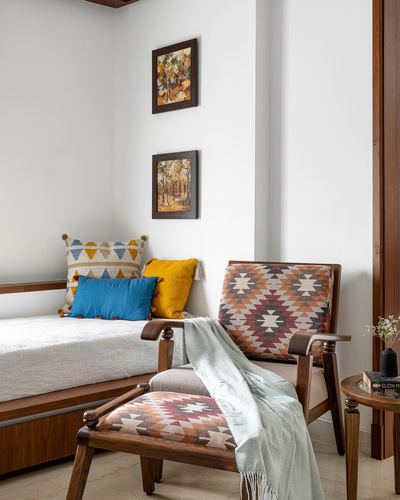 Wooden armchair with kilim pattern upholstery beside a daybed with colorful cushions and framed art