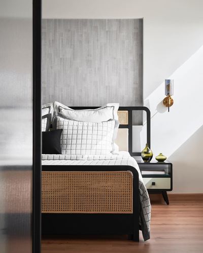 Bedroom with black cane bed, textured grey headboard wall, brass sconce, and sunlight cutting across the wall.