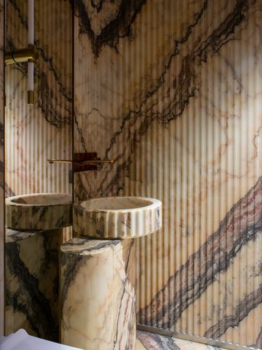 Fluted marble sink on marble pedestal against dramatically veined marble wall with brass fixtures