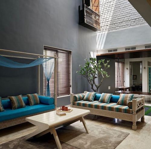 Carved wooden daybeds with teal cushions face a coffee table beneath a double-height grey courtyard wall
