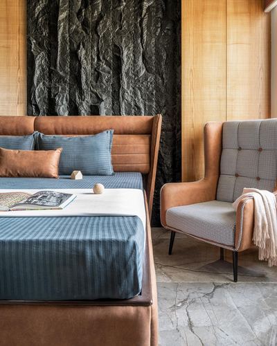 Bedroom with tan leather channel headboard, blue linens, houndstooth wingback chair, and stone feature wall