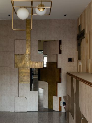 Brass-clad jigsaw-shaped door built into a textured plaster wall under a geometric brass pendant light