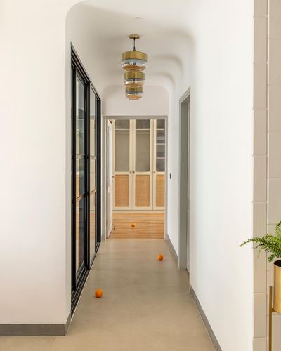 Arched white corridor with brass tiered pendant light, black glass door, oranges on floor, cane doors beyond
