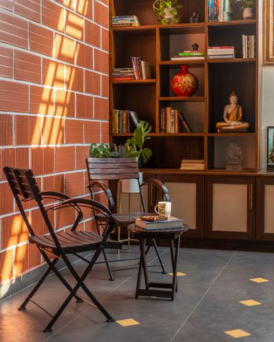 Sunlit corner with folding wooden chairs, round table, and bookshelf against a ribbed brick wall.