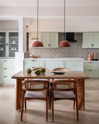 Wooden dining table with cane chairs facing sage green kitchen with patterned tile backsplash and terracotta pendants