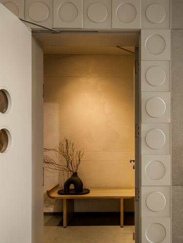 Open door with circular relief panels revealing a warmly lit alcove with wooden bench and vase