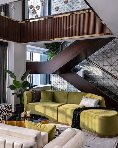 Olive velvet curved sofa beneath a dramatic zigzag wooden staircase with brass trim in a Mumbai penthouse