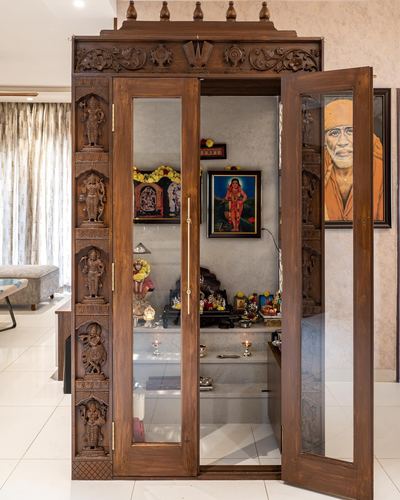 Ornately carved wooden pooja mandir with glass doors open revealing deity idols and lamps