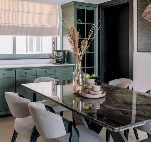 Dining table with dark stone top surrounded by grey chairs, sage green cabinetry behind