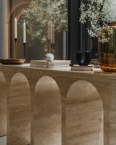 Travertine console table with arched base, styled with candles, books, and flowers near window