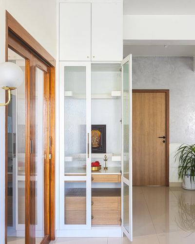 Built-in white pooja cabinet with ribbed glass doors, brass diya, and wooden door beside textured wall in Chennai apartment