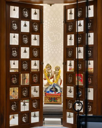Carved wooden doors with hanging bells open to a pooja alcove with deity idols under an om motif wall